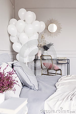 Bunch of white balloons in trendy bedroom interior with industrial dresser with golden mirror and bed with grey sheets Stock Photo