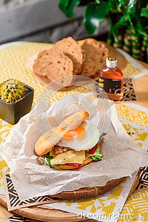 Burger with fried egg, chicken, cheese, tomatoes and other vegetables on a craft paper on yellow background Stock Photo