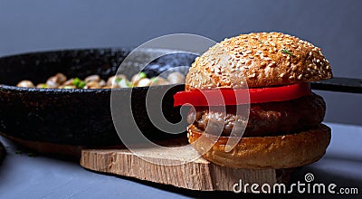Burger with a juicy cutlet tender bun and champignon mushrooms in a castiron pan on a wooden rustic plate on a gray background. Stock Photo