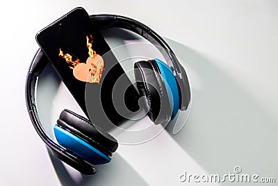 Burning heart symbol with burning flames on a black smartphone screen and blue over ear headphones and copy space Stock Photo