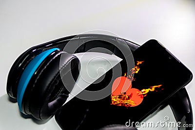 Burning heart symbol with burning flames on a black smartphone screen and blue over ear headphones and copy space Stock Photo