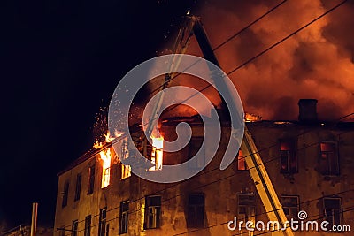 Burning house at night, roof of building in flames of fire and smoke, firefighter on crane extinguishes fire with water from hose Stock Photo