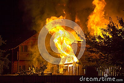 Burning wooden house at night. Bright orange flames and dense smoke from under the tiled roof on dark sky, trees silhouettes and r Stock Photo