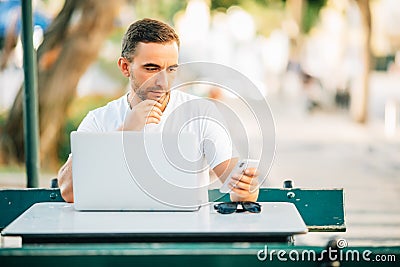 Business on fresh air. Handsome young man working on laptop and typing on the mobile phone while sitting at the wooden table outdo Stock Photo