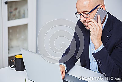 Business man teleworking from home with a laptop and talking on a cell phone. Bald man with glasses Stock Photo