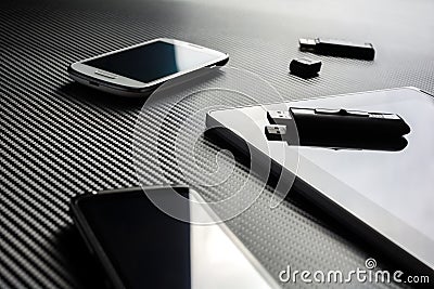 2 Business Mobiles With Reflections And An USB Drive Lying Next To A Blank Tablet With USB Drive On Top, All Above A Carbon Layer Stock Photo