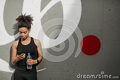 African american woman in sportswear with headphones, fitness tracker and bottle looks at smartphone Stock Photo