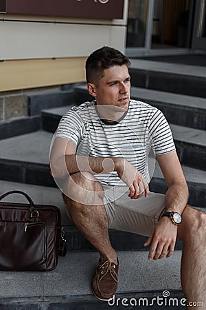 Business young man in a stylish summer striped T-shirt in fashionable shorts in suede shoes with a leather bag sits Stock Photo