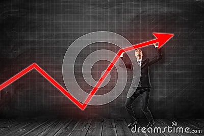 Businessman in black suit holding red arrow going up in room woth wooden floor near black graph ruled balckboard Stock Photo