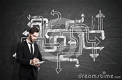 Businessman in a black suit with a notebook standing near a chalkboard with tangled arrows sketch on it. Stock Photo