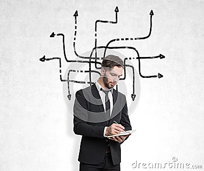 Businessman in a black suit with a notebook standing near a concrete wall with tangled arrows sketch on it. Stock Photo