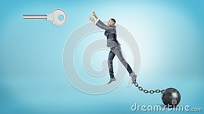 A businessman chained to an iron ball tries to jump and reach a large silver key hanging above. Stock Photo