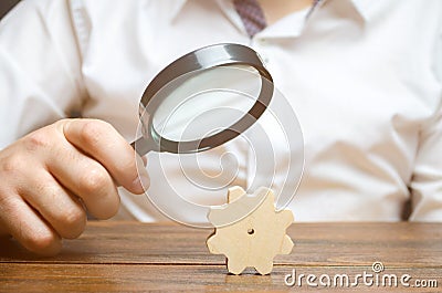 Businessman examines a wooden gear through a magnifying glass. study and analysis of business processes and subjects. Stock Photo