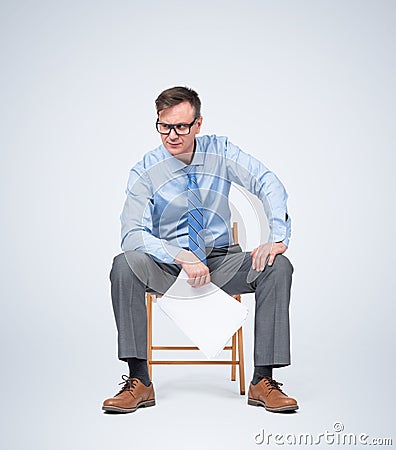 Businessman in glasses, a shirt and tie sits on a chair holding paper in his hands, on light blue background. Stock Photo