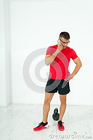 Businessman with glasses in shorts and t-shirt negotiates about business on his cell phone between kettle bell reps in the gym. Stock Photo