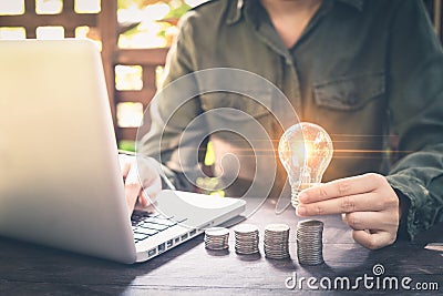 Businessman hand holding lightbulb on stack coins, and working with computer on workplace. Creative ideas concept of saving money Stock Photo