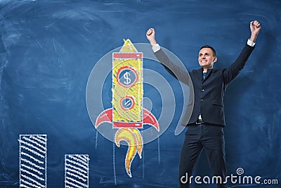 Businessman with hands raised in victory and rocket drawn in chalk on blue blackboard background Stock Photo