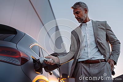 Businessman, holding power supply and charging his electric car during sunset. Concept of ecology tranport. Stock Photo