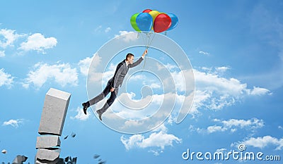 A businessman holding a set of colorful balloons on a string and flying away from a crumbling concrete column. Stock Photo