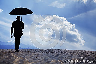 Businessman holding an umbrella and walking away in the middle of the desert with dreamlike clouds Stock Photo