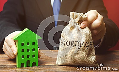 Businessman holds a money bag with the word Mortgage and a wooden house. Real estate concept. Loan for apartment and housing. Stock Photo