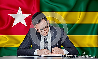 A businessman in a jacket and glasses sits at a table signs a contract against the background of a flag togo Stock Photo