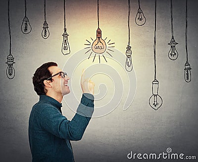 Businessman keeps index finger pointed up, eureka gesture, choosing a hanging glowing light bulb from others switched off. Stock Photo