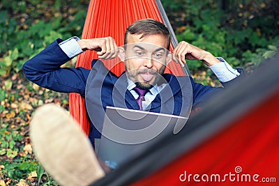 Businessman with laptop relaxs in a hammock on nature. He talking on video chat and doing funny faces with his tongue hanging out Stock Photo