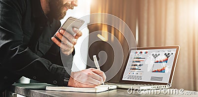 Businessman looks at laptop screen, making a note in notebook while holding smartphone.Entrepreneur analyzes information Stock Photo