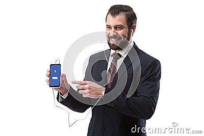 Cheerful bearded businessman listening music with earphones and pointing at smartphone with facebook website Editorial Stock Photo