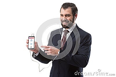 Cheerful bearded businessman listening music with earphones and pointing at smartphone with instagram website Editorial Stock Photo