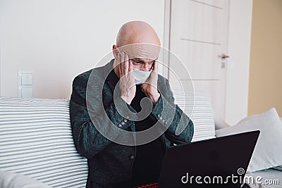Businessman in protective mask sitting on the sofa he sees terrifying stock market results on the laptop. Stock Photo