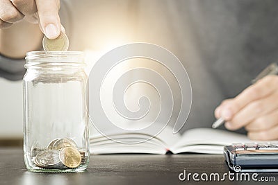 Businessman putting coin into jar glass and record on notebook. Stock Photo