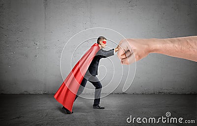 A businessman in a red cape and a mask pushing at a giant human fist on concrete background. Stock Photo