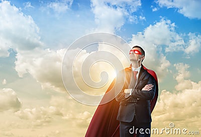 Businessman in a red cape and a red eye mask standing with hands crossed on clouded sky background. Stock Photo