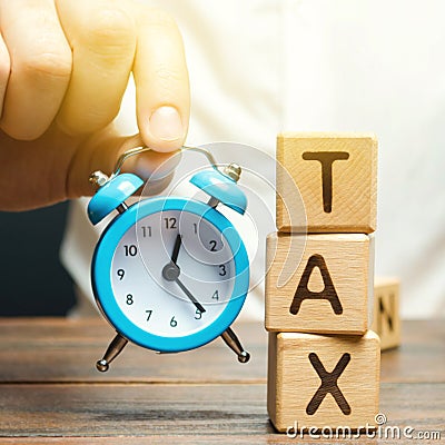 Businessman`s hand holding a clock near the wooden blocks with the word Tax. Time to pay taxes. The concept of annual taxation. Stock Photo