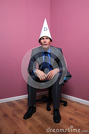 Businessman sat in corner wearing dunce hat Stock Photo