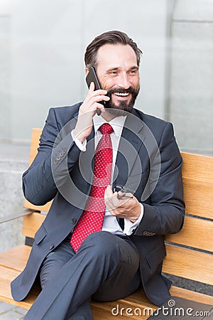 Businessman sitting on bench using his cell phone. Smiling bearded businessman sitting on bench and talking on the phone Stock Photo