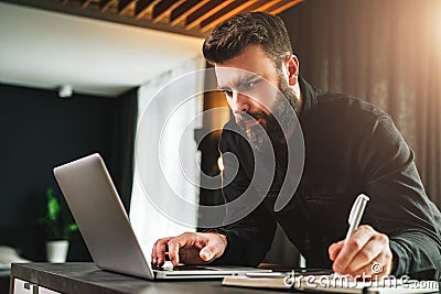 Businessman is standing by computer, looking at laptop screen, making notes in notebook. Man watching webinar, learning. Stock Photo