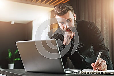 Businessman is standing near computer, working on laptop, making notes in notebook. Man watching webinar, learning. Stock Photo