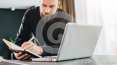 Businessman is standing near computer, working on laptop, making notes in notebook. Man watching webinar, learning. Stock Photo