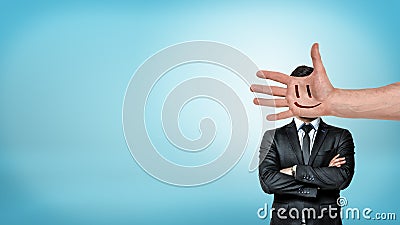 A businessman stands in a front view with folded arms behind a giant male hand with a smiley face covering his head. Stock Photo