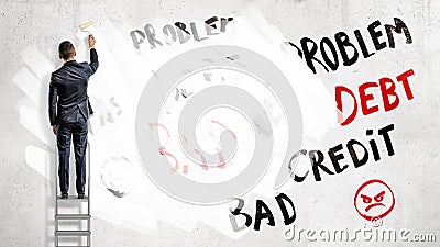 A businessman stands on a step ladder and paints over words Problem, Debt and Credit with a white paint roller. Stock Photo