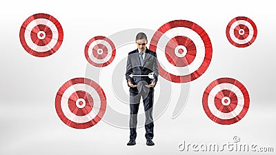 A businessman stands beside a wall with many archery targets and looks at an arrow hovering over his hands. Stock Photo