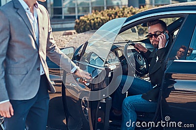 Businessman in sunglasses is talking by mobile phone while his young smart assistant is opening door for him. Stock Photo