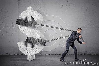 A businessman tugs at a chain trying to move a large concrete dollar sign from its place. Stock Photo