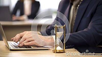 Businessman typing on laptop at desk, hourglass trickling, deadline approaching Stock Photo