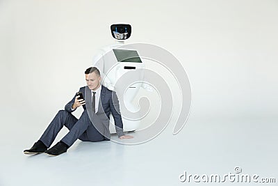 Businessman uses a smartphone while sitting on the floor next to a robot. Modern Robotic Technologies. Humanoid Stock Photo