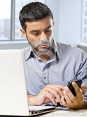 Businessman working on computer laptop using mobile phone at office desk in front of skyscraper window Stock Photo