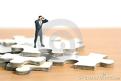 Businessmen standing above jigsaw puzzle piece stack using binoculars above wooden table. Miniature tiny people toys photography Stock Photo